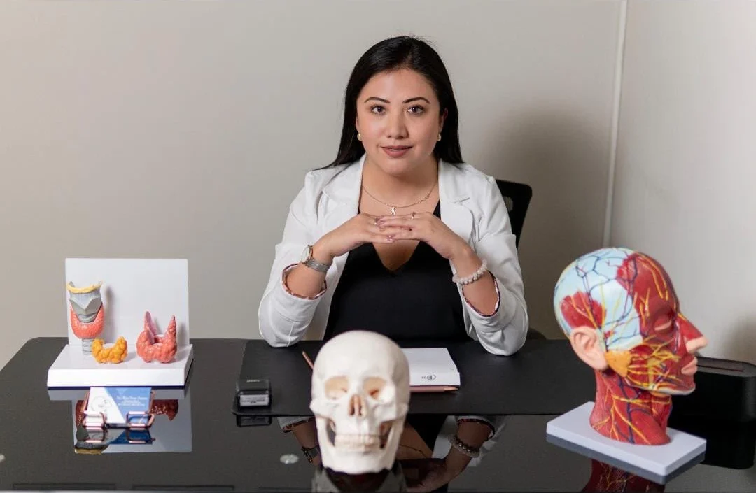 Dra. Alytsa Garcia sentada en su escritorio con modelos anatómicos de tiroides, cráneo y cabeza y cuello